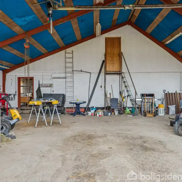 Garage med redskaber og værktøj spredt rundt. Arbejdsborde står i midten, og der hænger stiger på væggen. Loftet er båret af træbjælker, miljøet virker travlt og praktisk.
