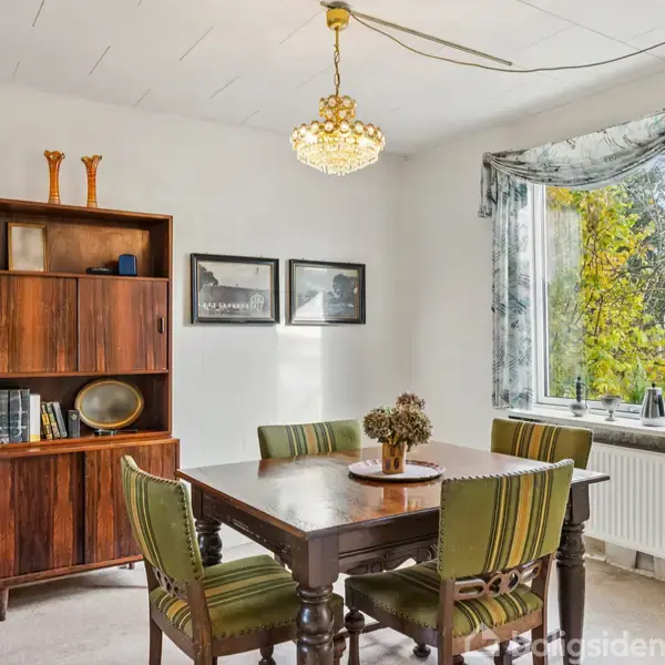 Et træbord med fire grønne stole står i en spisestue. En bogreol med bøger og vaser er langs væggen. Et vindue med blomstermønster-gardiner og et lysstofrør ses ved siden.