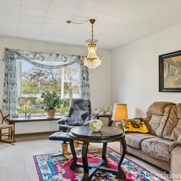 Stue med lænestol og sofa placeret omkring et rundt bord. Lysekrone hænger fra loftet. Foto og lampe er på væggene, og stort vindue giver udsigt til haven.