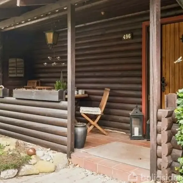Træhus med terrasse-område, hvor et træbord og en stol er placeret. Husnummeret "496" hænger ved døren. Omgivet af grønne buske og stier af sten.