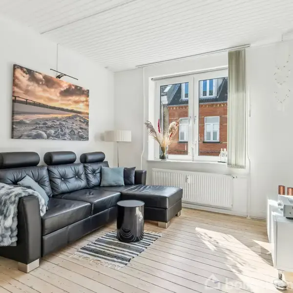 Sort lædersofa står i en lys stue med trægulv, flankeret af et sofabord og reol. Et stort vindue lader sollys strømme ind, med udsigt til murstensbygning.