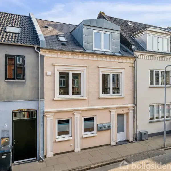 Et to-etagers byhus med lyserød facade og hvide vinduer står på en rolig gade. Huset har tagvinduer og er omgivet af tilstødende bygninger i forskellige farver.