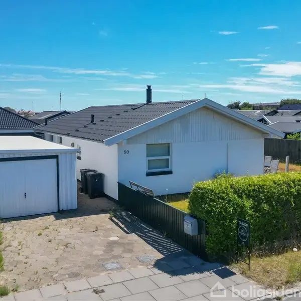 En hvid bungalow med fladt tag står ved siden af en garage. Husnummeret "50" ses på facaden, omgivet af en grøn hæk og beliggende i et villakvarter under en klar blå himmel.