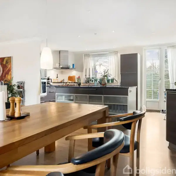 Køkken-alrum med træbord og stole i forgrunden. Køkkenet i baggrunden har moderne skabe og apparater. Store glasdøre giver naturligt lys. Dekorative elementer og planter pryder rummet.