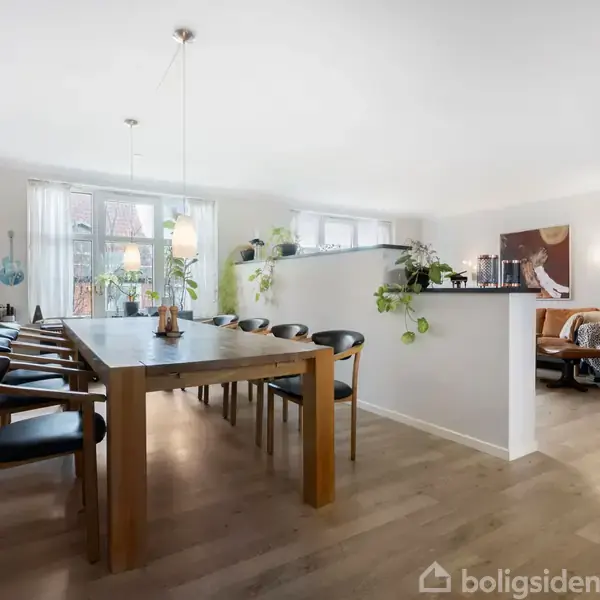 Spisebord med otte stole står i lys spisestue med trægulv. Lys strømmer ind gennem store vinduer. Ved siden af er en hyggekrog med lænestole og planter.