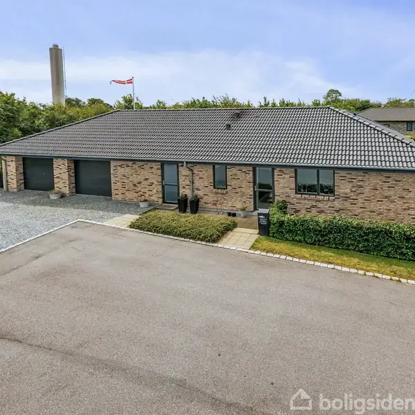 Et moderne murstenshus med gråt tegltag står i rolig kvarter. To sorte garageporte på venstre side, grusbelagt indkørsel, grønne buske foran huset og en flagstang i baggrunden.