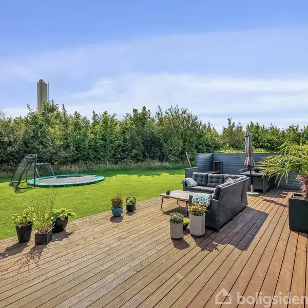 Terrasse med udendørsmøbler, mange blomsterpotter og en parasol står på trædæk. Græsplænen har en trampolin, flagstang og et omringende hegn af buske.