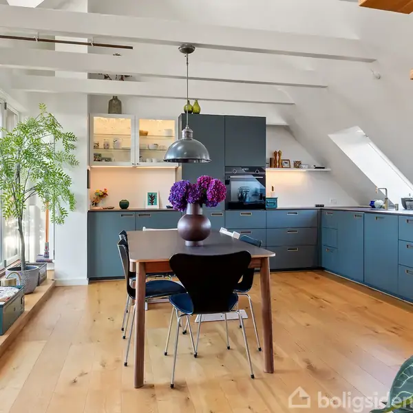 Et træbord med lilla blomster i vase står i et moderne køkken med blå skabe. Store vinduer og et ovenlys tilføjer naturligt lys. Planter dekorerer rummet.