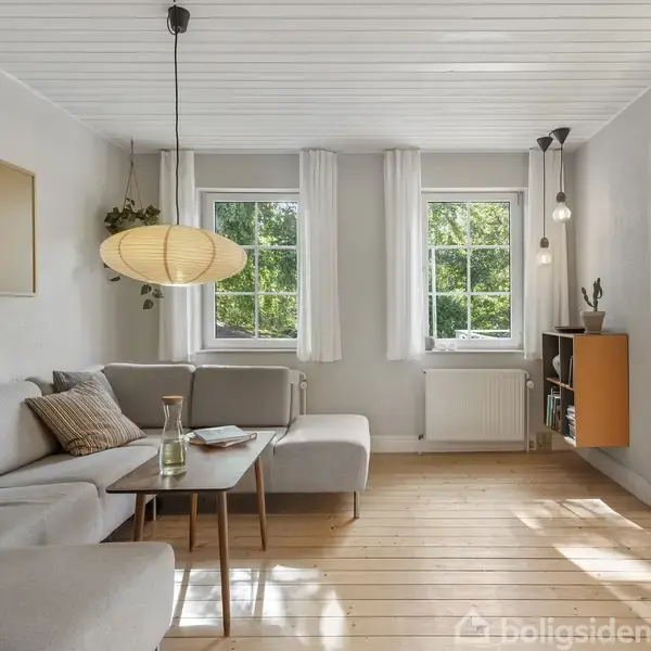En lys stue: Grå hjørnesofa står tæt på træbord, mens en lampe hænger over den. Store vinduer og hvidt loft skaber en lys atmosfære. Trægulv og grøn udsigt udenfor.
