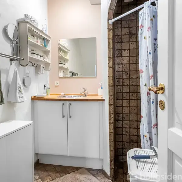 Badeværelse med hvidt møbel under spejl, bruser med blomsterforhæng til højre, og håndklæder hænger på en stang. Vægge er lyse, og der er fliser på gulvet.