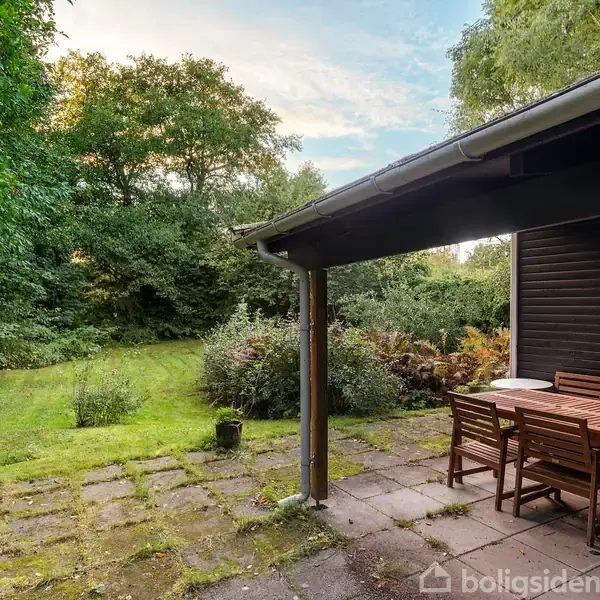 Terrassemøbler står på en flisebelagt patio ved et hus. Omkring er der frodigt grønt græs og buske, og træer omslutter stedet, hvilket skaber en afsondret atmosfære.