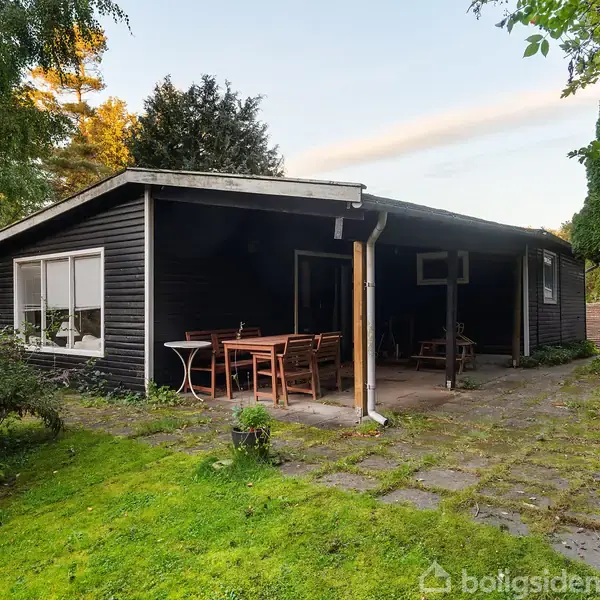 Sort træhus med en overdækket terrasse, hvor et træbord og stole står placeret. Den omgivende have er frodig med grønne planter og træer.