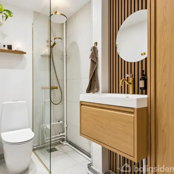 Badeværelse med toilet og vask. Vasken har et rundt spejl og en sæbedispenser. Til venstre er bruseniche med glasvæg. Grøn plante hænger fra loftet på en hylde.