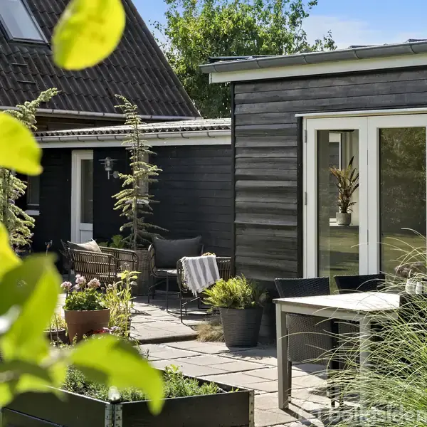 En moderne træbygning med store vinduer står ved siden af en lille terrasse med borde og stole. Haven er fyldt med grønne planter, og der er en blå himmel i baggrunden.