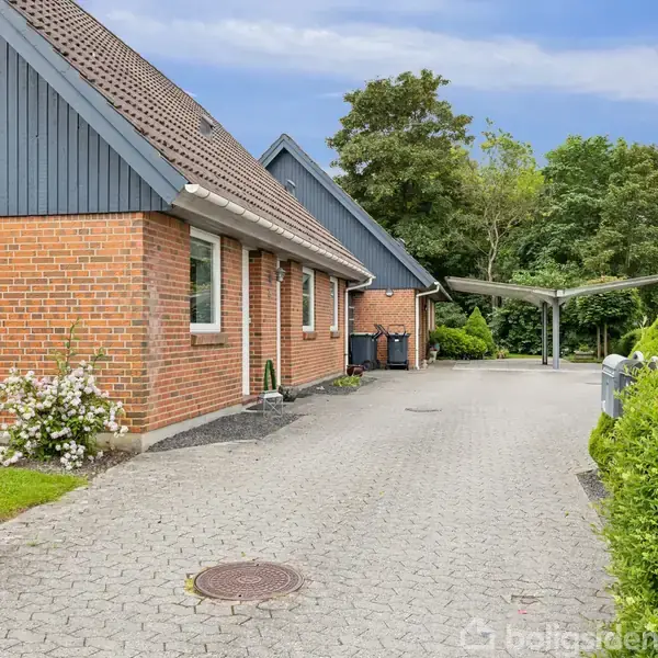 Rødt murstenshus med blå gavle står roligt i en villahave. En flisebelagt indkørsel leder til en carport. Buske og træer omgiver området, der er fyldt med naturlig grønne.