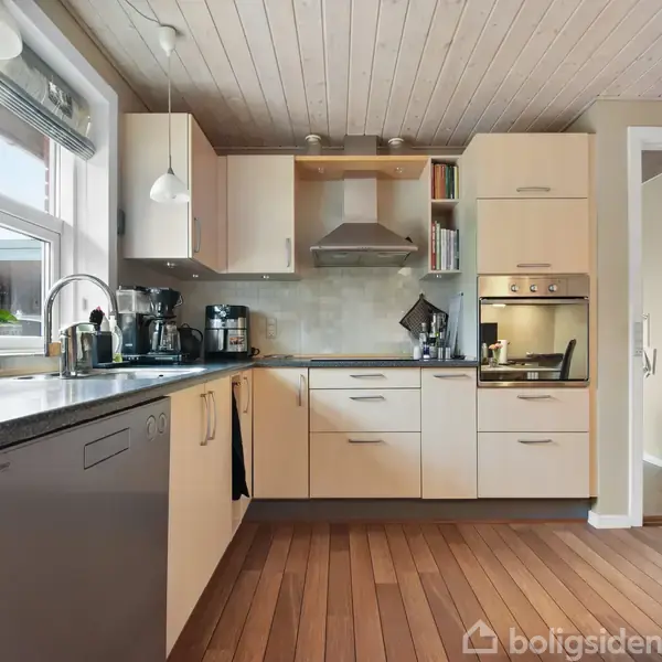 Køkken med lyse skabe og trægulv, udstyret med opvaskemaskine, kaffemaskine og ovn, set i dagslys gennem et stort vindue. En plante står ved siden af vasken.