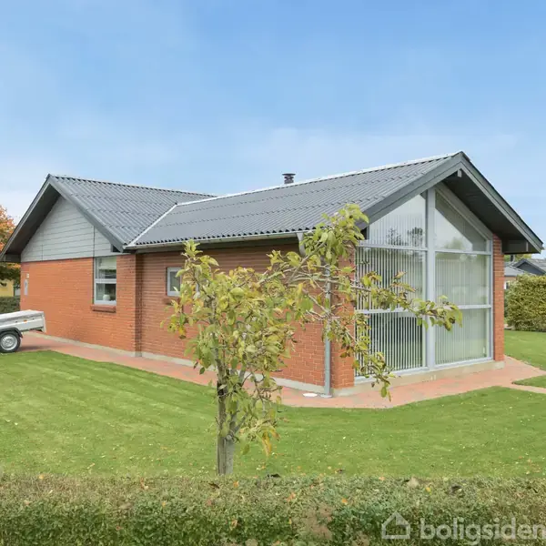 Rødt murstenshus med moderne design står på en velplejet græsplæne. Glasfacade på fronten og sort tag. Lille trætrailer ved siden af. Blomstrende træer omgiver gårdspladsen.