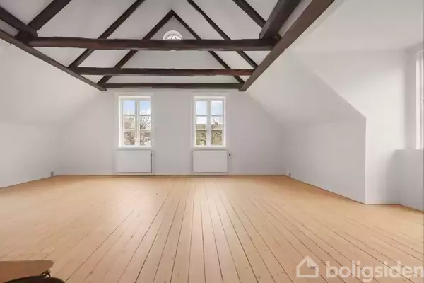 Et rum med skrålofter og synlige træbjælker står tomt. Trægulvet er lyst, og to vinduer tillader naturligt lys at strømme ind udefra.