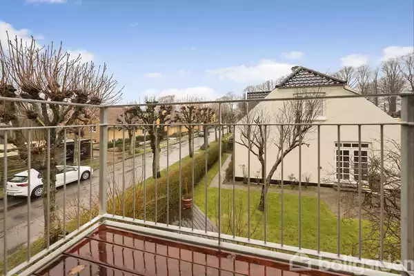 Et altanområde med vådt gulv, set fra anden sal, vender mod en vej med parkerede biler og træer foran en hvid bygning med græsplæne.