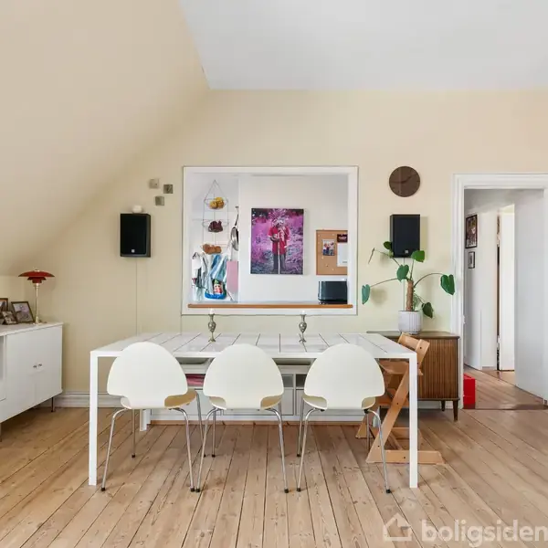 Et lyst rum med et spisebord og tre hvide stole i midten, omgivet af lyst træ, en radiator til højre, indrammede billeder og planter på møbler.