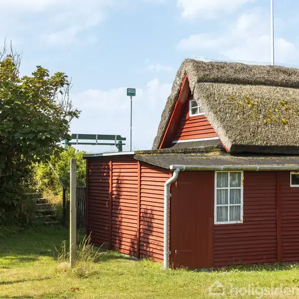 Et rødt træhus med stråtag står stille i en grøn have omgivet af buske og træer. Et metalhegn og en frafløjet savskivelygte kan ses i baggrunden.
