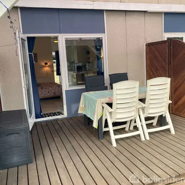 Udendørs terrasse med hvidt plastikbord og stole, dækket med en farverig dug. I baggrunden ses en åben dør, der fører ind til et værelse med en opredt seng.