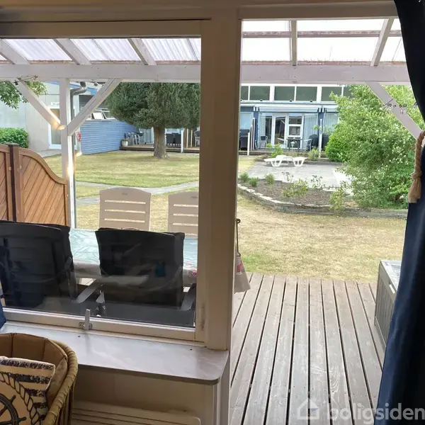 Vindue åbner ud til en træterrasse med spisebord og stole. I baggrunden ses en græsplæne, en rund cirkel af planter og et hus med store vinduer.