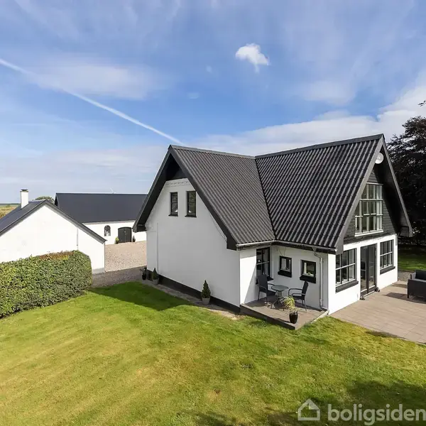 Et hvidt hus med sort tag står ved en terrasse med havemøbler, omgivet af grøn græsplæne og buske. En mindre bygning ses i baggrunden under en blå himmel med nogle skyer.
