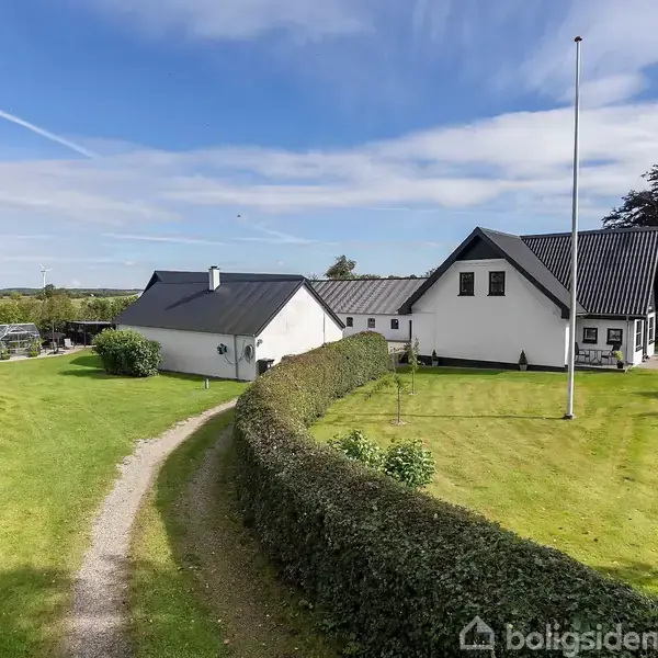 En hvid villa med sort tag står i en frodig have. En sti snoer sig gennem den grønne græsplæne, omgivet af en tæt klippet hæk.