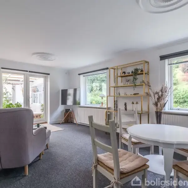 En stue med grå stol ved siden af en hvid sofa, et rundt spisebord og stole. Et vindue viser en have udenfor. En reol med pynt står i hjørnet.