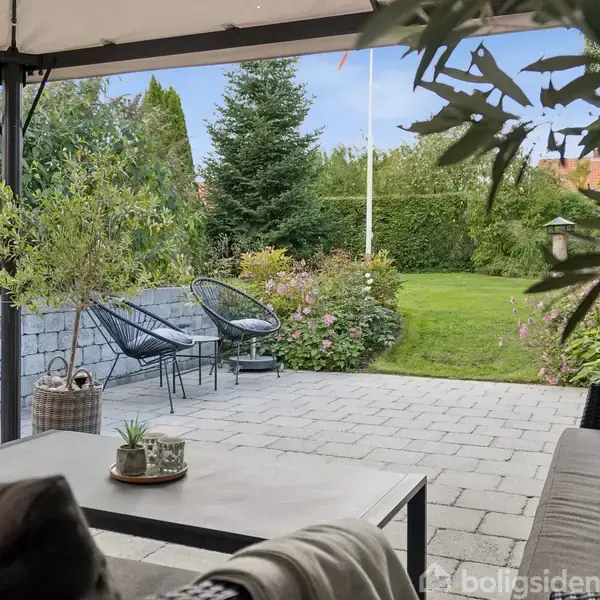 Terrassemøbler står under en pavillon med udsigt til en velholdt have. To moderne stole står ved siden af tæt bevoksning. En havegang fører til en velplejet græsplæne omkranset af blomstrende buske.