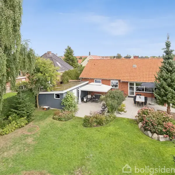 Rødt murstenshus står stille i en velplejet have med grønne græsarealer og forskellige træer; en flisebelagt terrasse med havemøbler ses ved siden af huset.