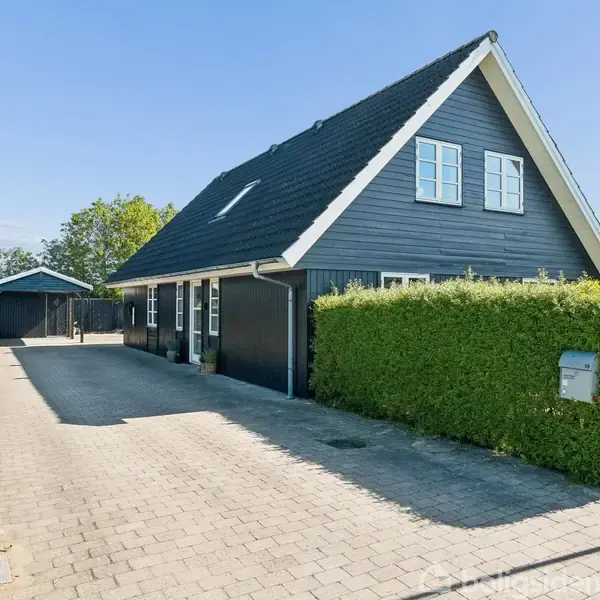 Et sort-hvidt hus står på en flisebelagt indkørsel med en carport i baggrunden. Hækken omgiver ejendommen under en klar blå himmel med grønne træer i nærheden.