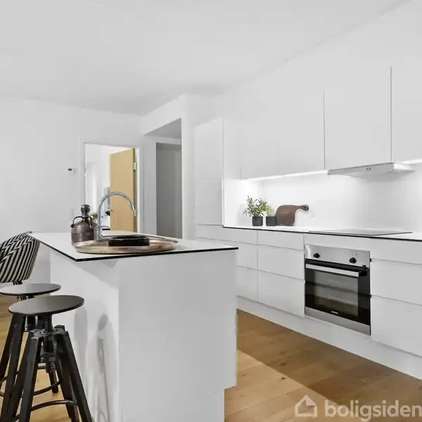 Et moderne køkken med hvide skabe og indbygget ovn. Køkkenø med træbarstole. I baggrunden ses et spisebord med stribede stole og en hængende lampe.