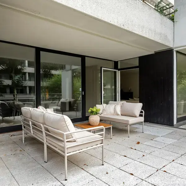 To patio med hvide sofaer og et bord, hvor der står en plante. Store glasfacader lukker lyset ind i en moderne bygning. Udenfor er der grønt omkring.