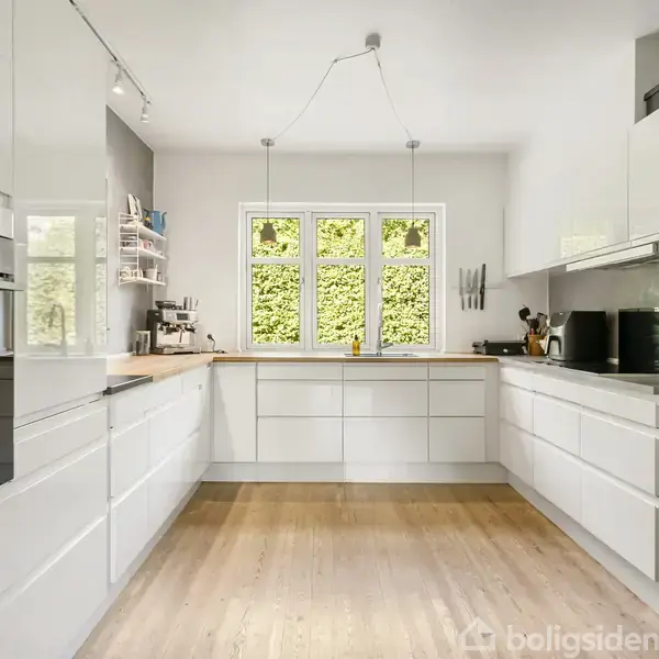 Køkken med hvide skabe og moderne apparater. Køkkenet er velindrettet med træbordplader og store vinduer med udsigt til en grøn baggrund, hvilket giver et lyst og rummeligt indtryk.