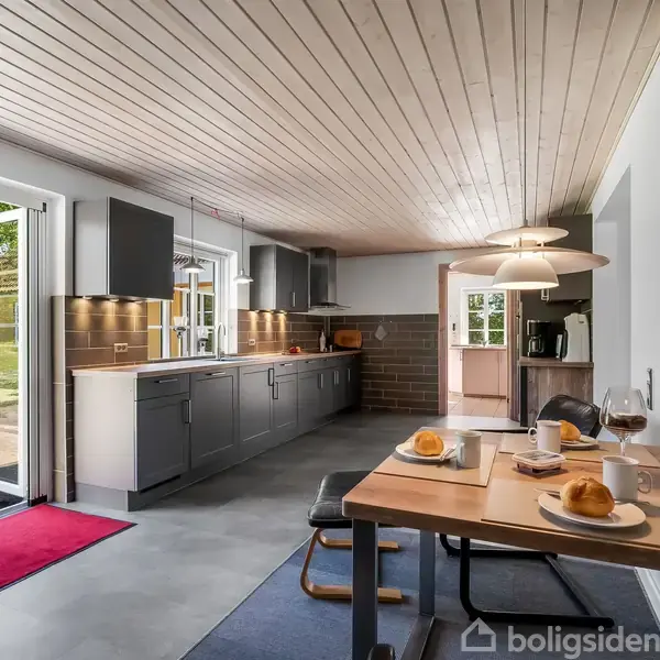 Et stilfuldt køkken-alrum med en træbordplade, morgenmad serveret med juice, croissanter og glas. Dør fører ud til en terrasse med udsigt til grønne træer.