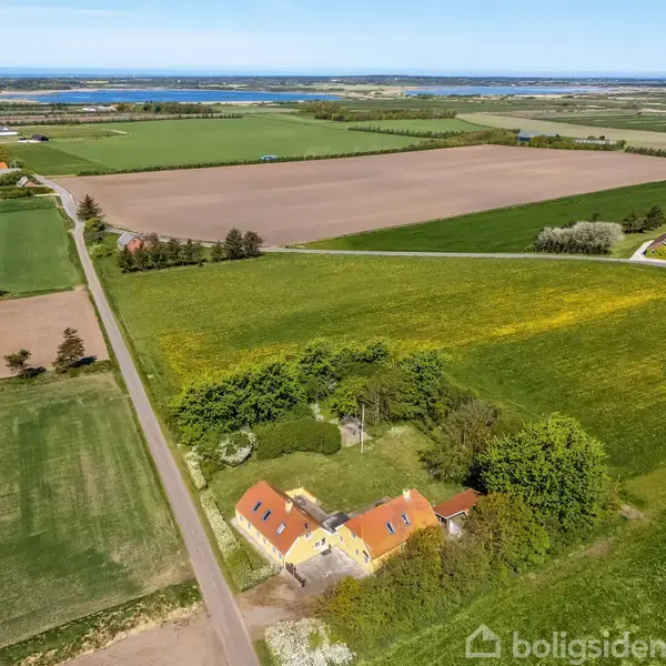 Et gult hus ligger blandt træer tæt ved en vej, omgivet af marker i forskellige grønne og brune nuancer, med udsigt til en stor sø i horisonten.