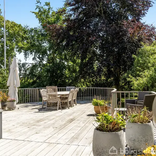 En stor træterrasse med havemøbler, parasol og blomsterkrukker. Terrassen omkranses af en lav, hvid rækværk med grønne træer og buske i baggrunden, der skaber en frodig atmosfære.