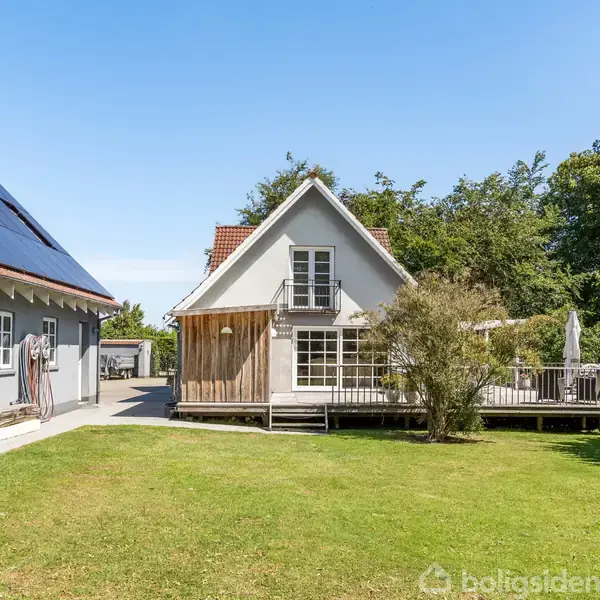 Et hvidt hus med en veranda står stille i en frodig have. Solpaneler er placeret på et tilstødende gråt udhus, omringet af grønne træer og blå himmel.