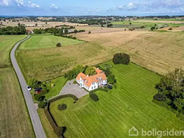 En hvid villa med orange tag står på en stor, velplejet græsplæne omgivet af marker og en snoet vej i landlige omgivelser under en blå himmel med skyer.