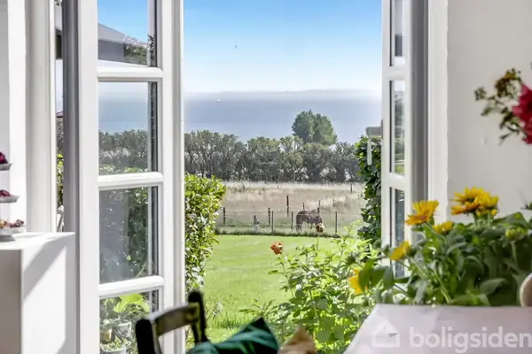 Et åbent vindue viser en udsigt over en frodig have med blomster og en mark med en hest. Træer og havet er synlige i baggrunden under en klar blå himmel.