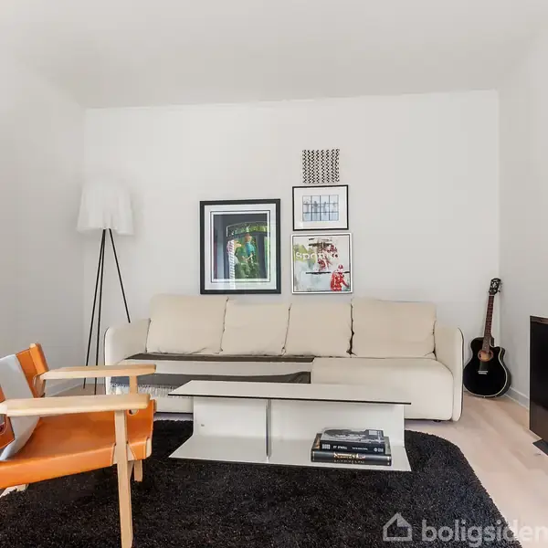 En lys stue med en beige sofa placeret mod en hvid væg med tre hængende billeder. Et sofabord står på et sort tæppe, omgivet af en orange lænestol, en stående lampe og en guitar i hjørnet. En fladskærms-tv står til højre.