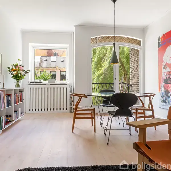 Et lyst, moderne rum med spisebord og stole i centrum. Til venstre står en bogreol, og der er farverig kunst på væggen. Store vinduer åbner ud mod en balkon.