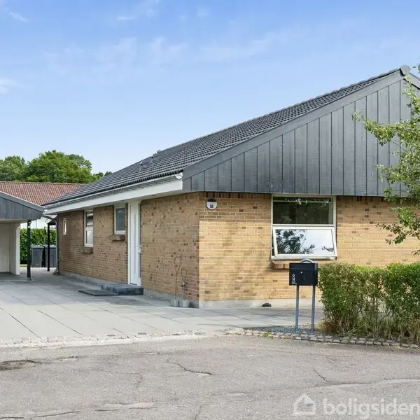 Et murstenshus med skråt tag står stille i et villakvarter. Der er en tilhørende garage til venstre og en flisebelagt indkørsel omgivet af grønne buske.