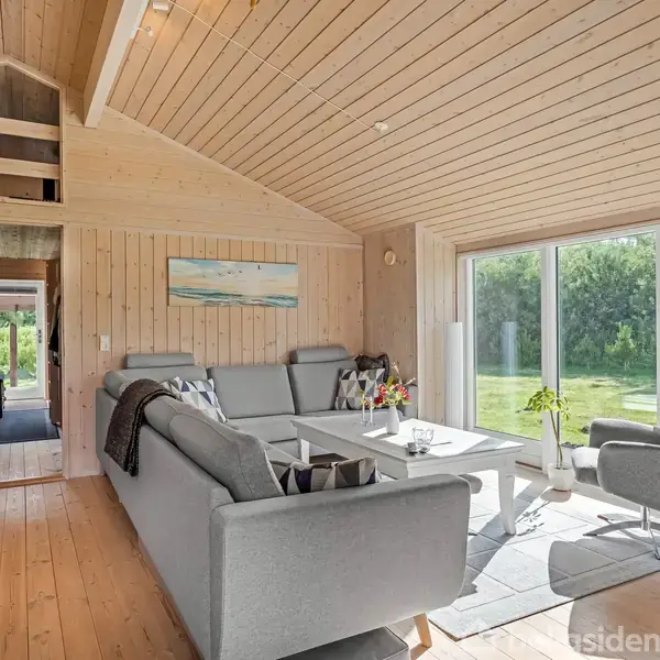 Et opholdsrum med grå sofaer og et sofabord står i sollys foran store vinduer. Trævægge og -lofter skaber en hyggelig atmosfære. Gennem en åben dør ses et entréområde.