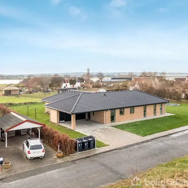 Et moderne køkken-alrum med åbent rum. En spiseplads har sorte stole omkring et bord, mens et køkkenområde har en kogeø med barstole. Overalt er der lyse vægge og lofter med indbyggede spotlights. Blomster og kunst pryder området.