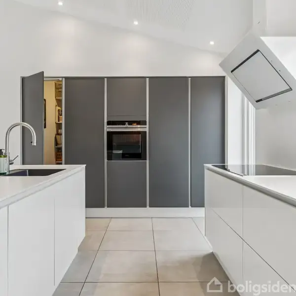 Moderne køkken med hvide skabe og en central ø med indbygget vask og armatur. Grå skabsvæg med integreret ovn og keramisk kogeplade under emhætte. Lys, minimalistisk atmosfære.