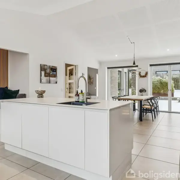 Køkken med hvid ø i midten, udstyret med vask og vandhane. I baggrunden ses spisebord, stole og store vinduer, der åbner mod en terrasse med haveudsigt.