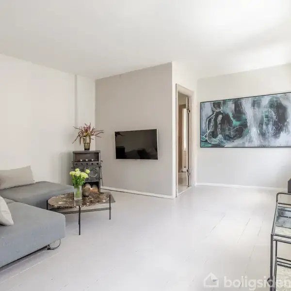 En moderne stue med en grå sofa og sofabord med blomster. En vægmonteret TV hænger foran sofaen. Kunst maleri på væggen; lys kommer ind fra højre vinduer.